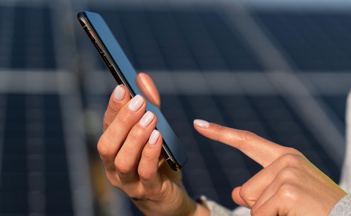 Zwei Hände mit einem Smartphone, im Hintergrund sind Solarmodule